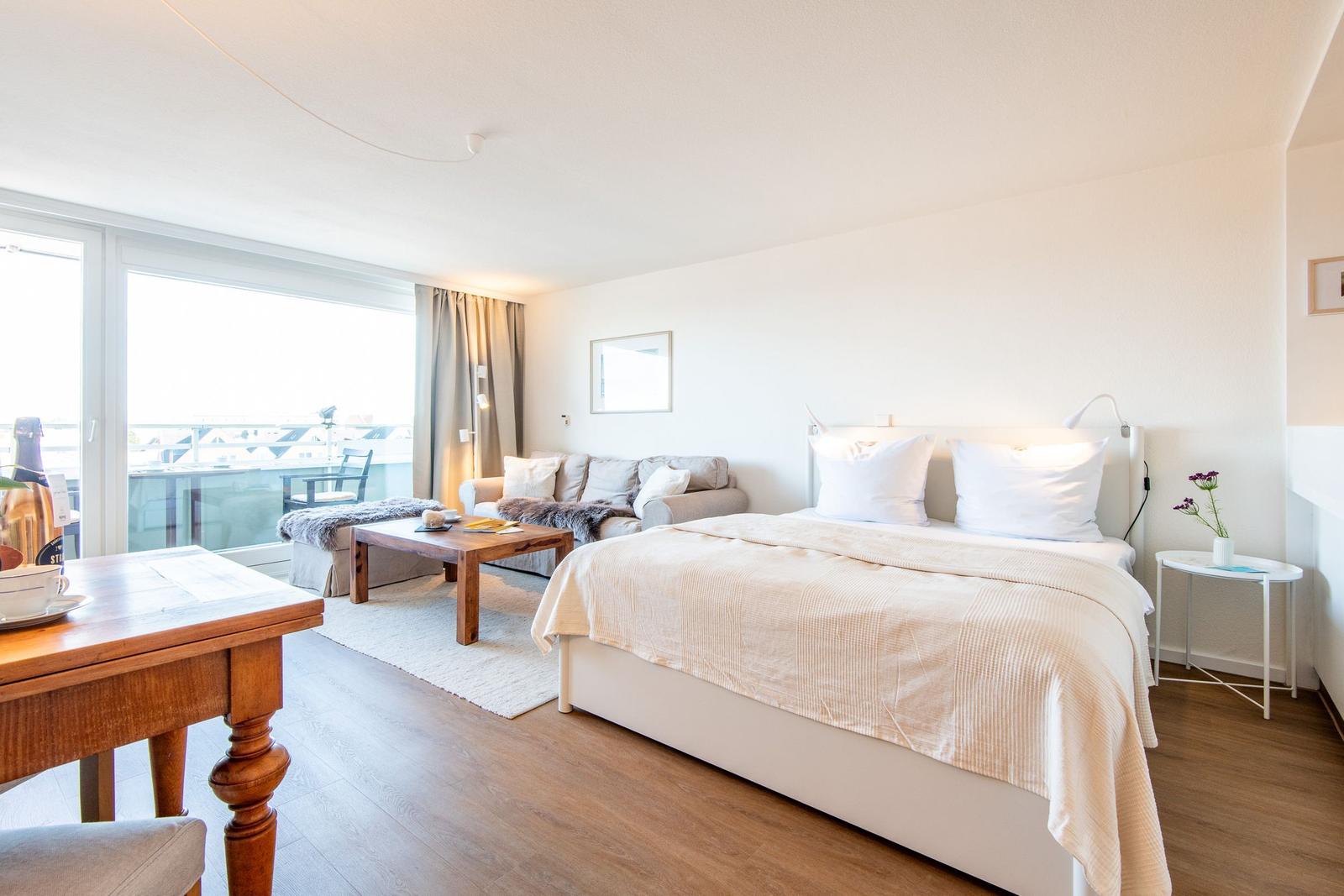 Ein Schlafzimmer mit Bett, Sofa, Tisch und Balkon. Holzfußboden und helle Wände.