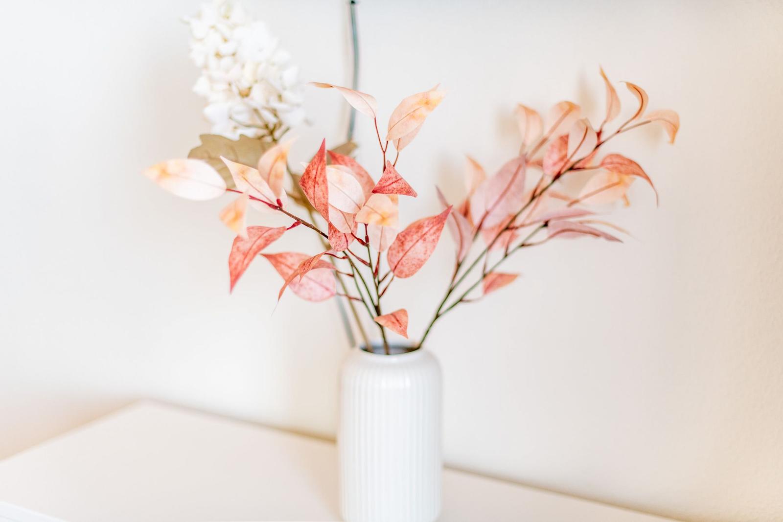 Ein weißer Vase mit Blättern und Blüten auf einer weißen Oberfläche.