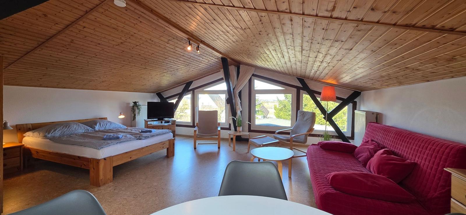 Schlafzimmer mit Doppelbett, Sofa und großem Fenster. Holzdecke und Parkettboden.