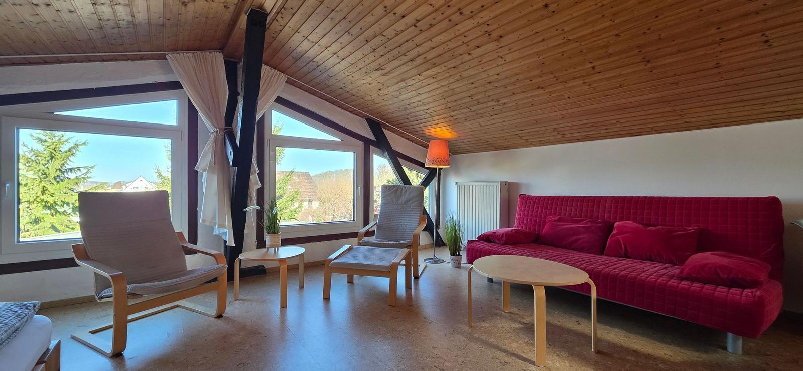 Wohnzimmer mit rotem Sofa, zwei Sesseln und großen Fenstern. Holzdecke und Boden.