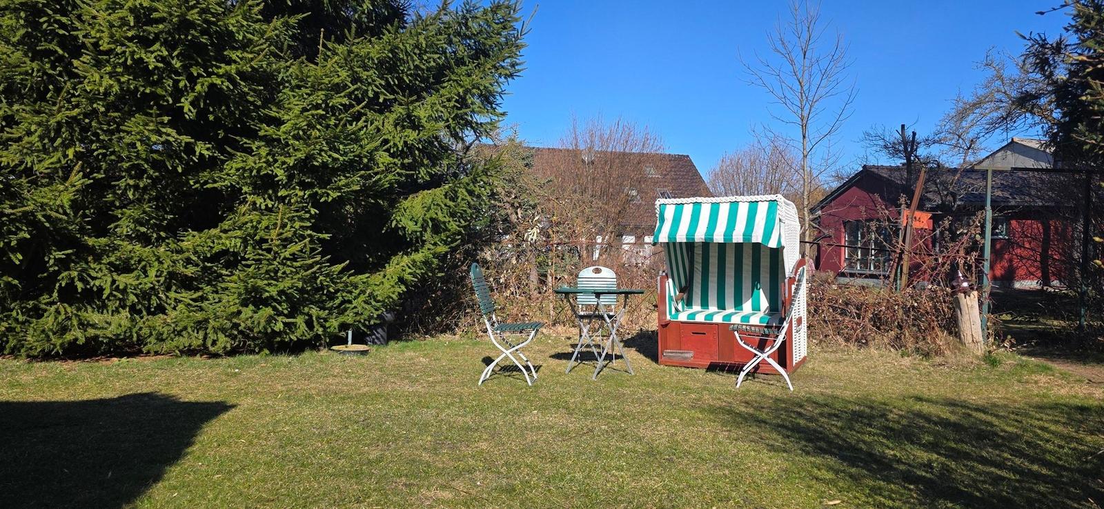 Garten mit Strandkorb, Tisch und Stühlen. Grasfläche, Bäume und Hintergrundhäuser.