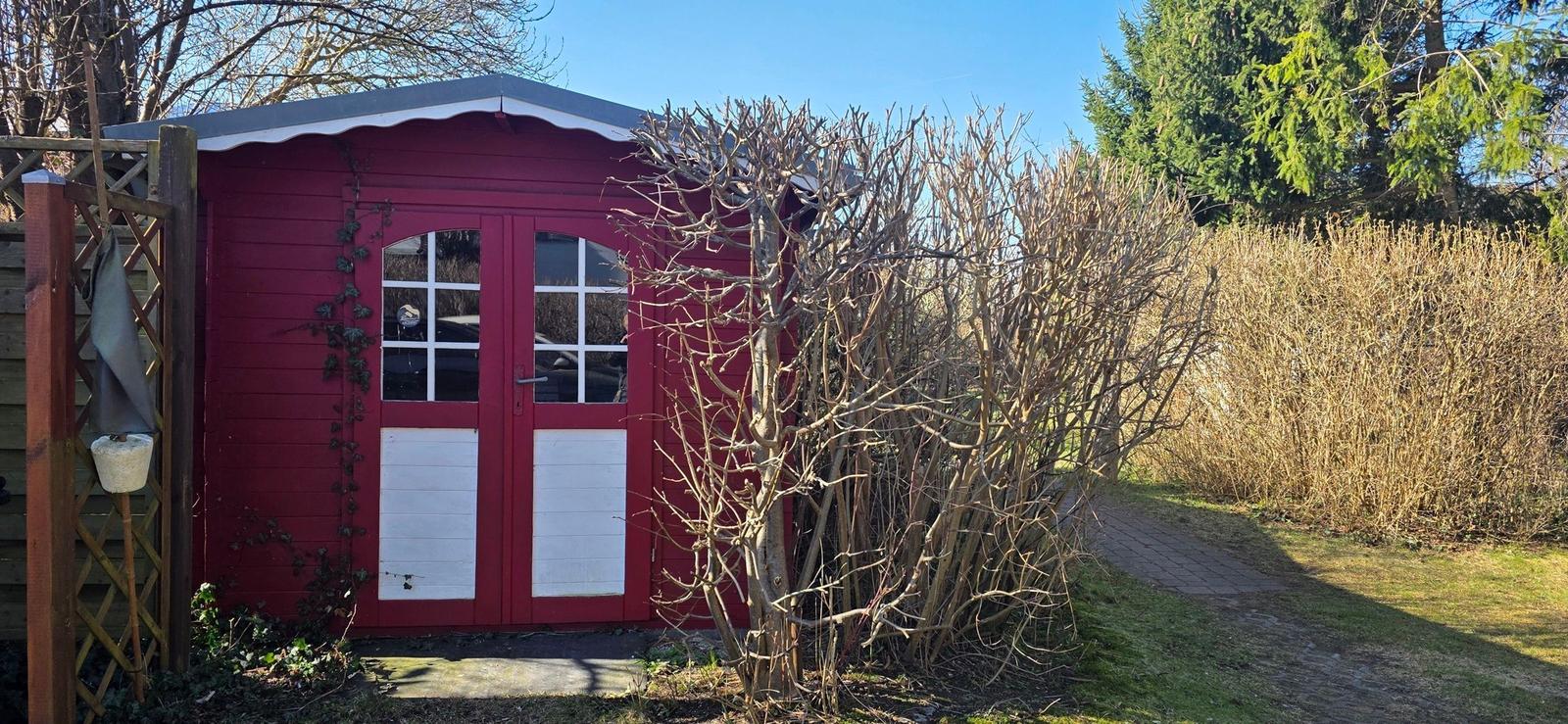Roter Gartenhaus mit Fenster und Pflanzen im Hintergrund.
