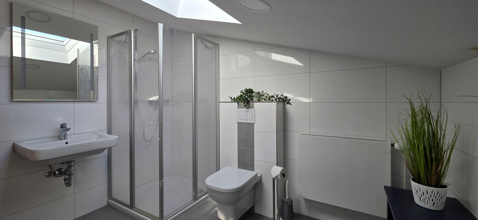 Badezimmer mit Dusche, Waschbecken und Toilette. Weiße Fliesen und Pflanzen.