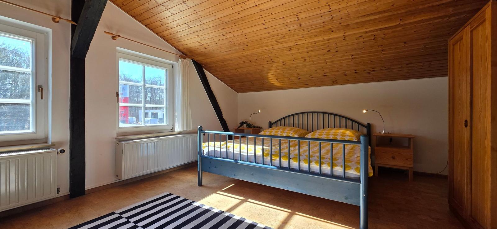 Schlafzimmer mit Holzdecke, Bett mit gelbem Bettbezug und zwei Fenstern.