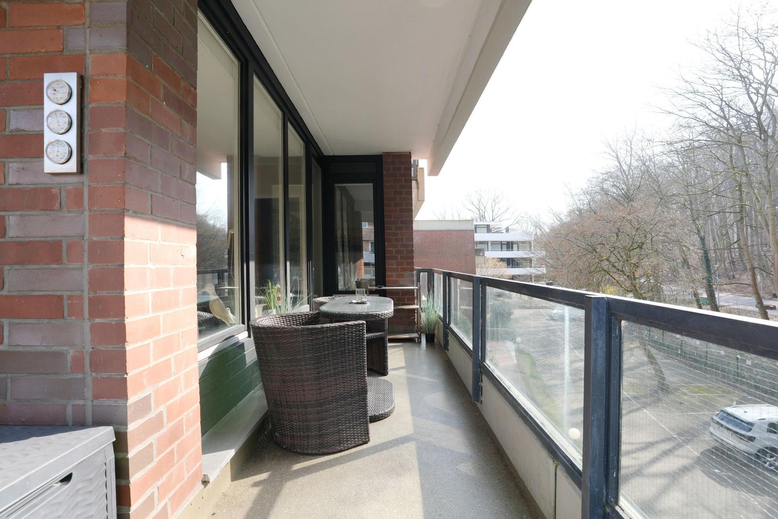 Balcony with wicker furniture and glass railing overlooking trees and parking area.