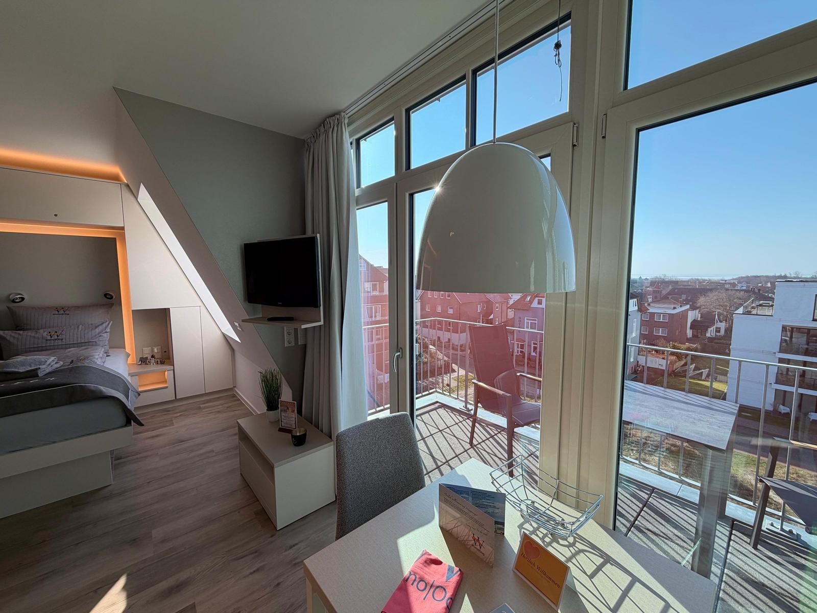 Schlafzimmer mit Balkon und TV. Große Fenster bieten Blick auf die Stadt.