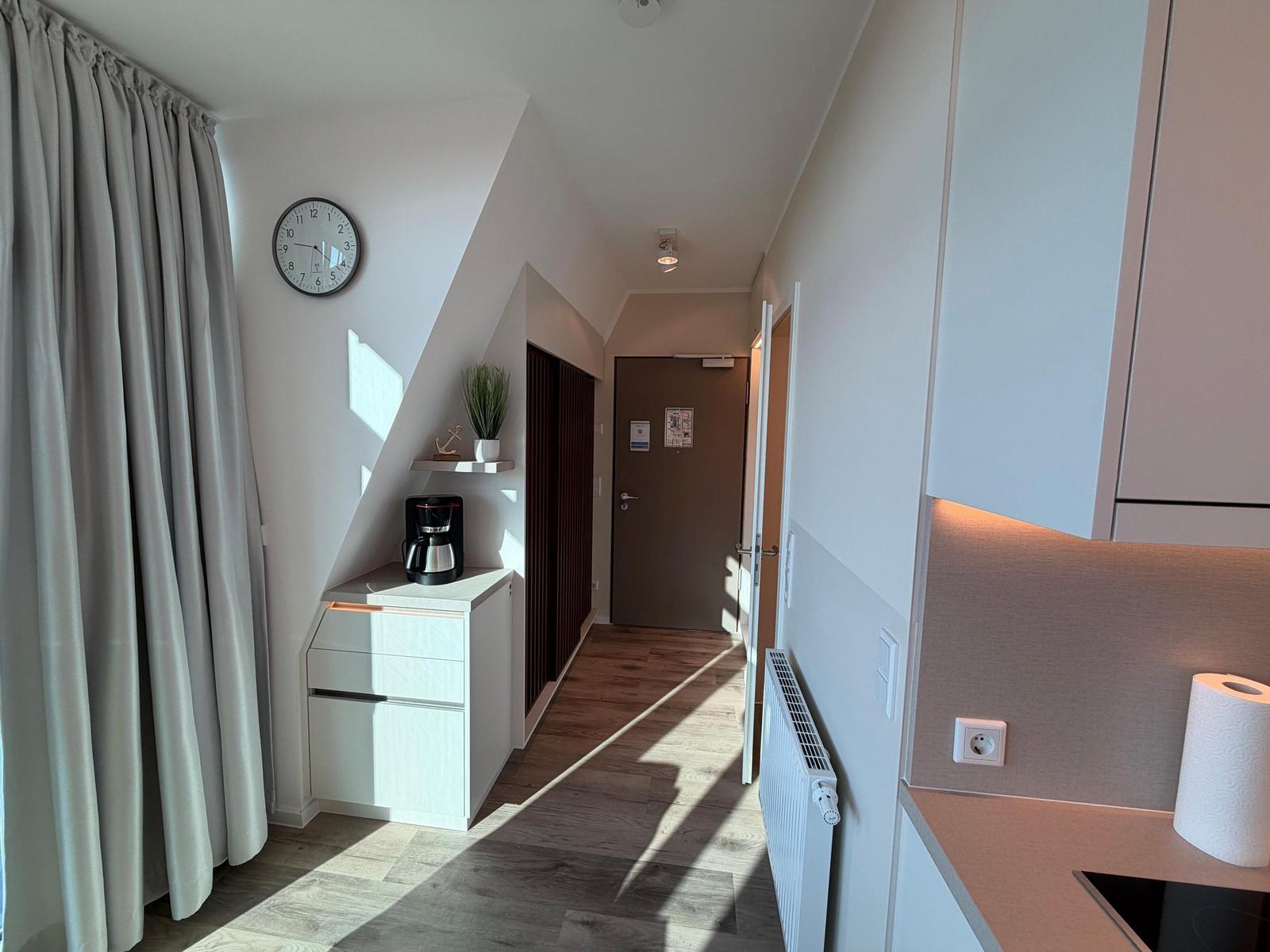 Flur mit Küchenzeile, Kaffeemaschine und Wanduhr. Holzfußboden und Fenster mit Vorhang.