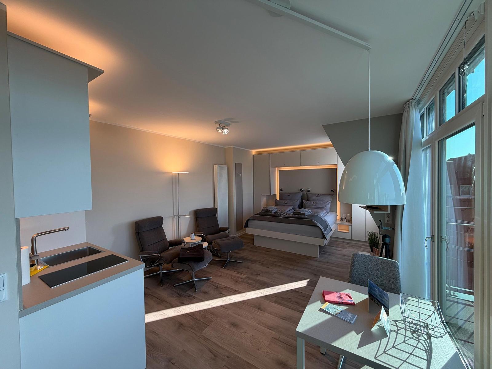 Schlafzimmer mit Bett, Sitzgelegenheit und Küchenbereich. Große Fenster und moderne Einrichtung.