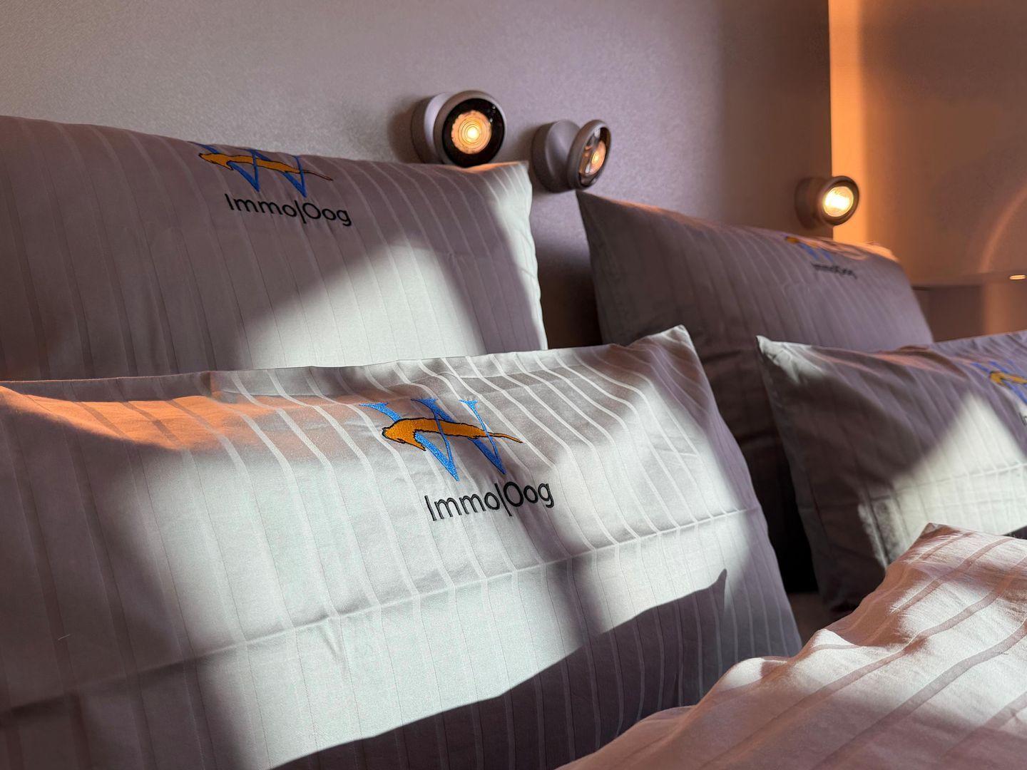 Bettkopfteil mit Kissen und Wandlampen. Kissen mit Logo.