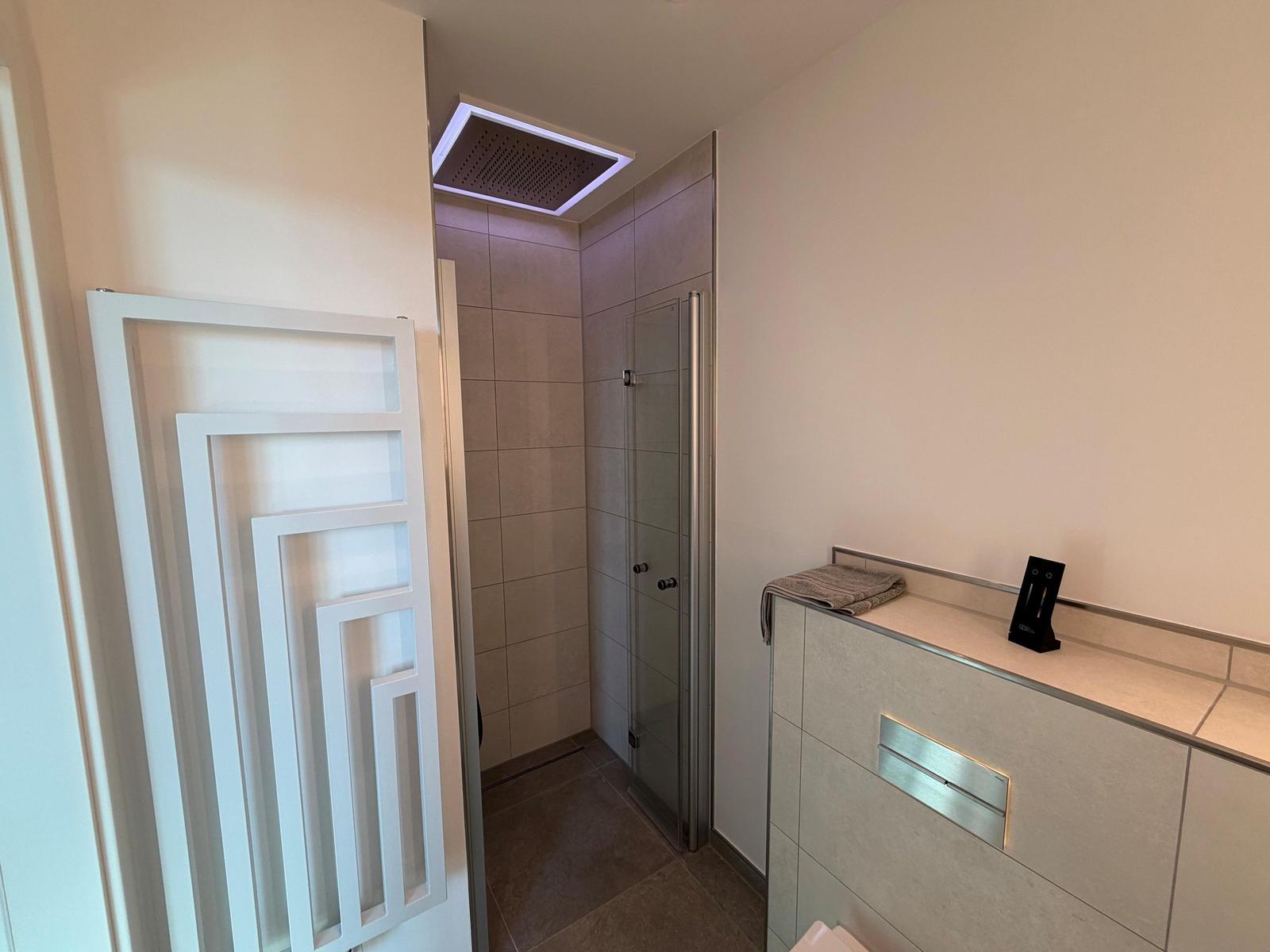 Moderne Dusche mit Glaswand und Deckenbrause. Neben einem Waschbecken mit Handtuch.