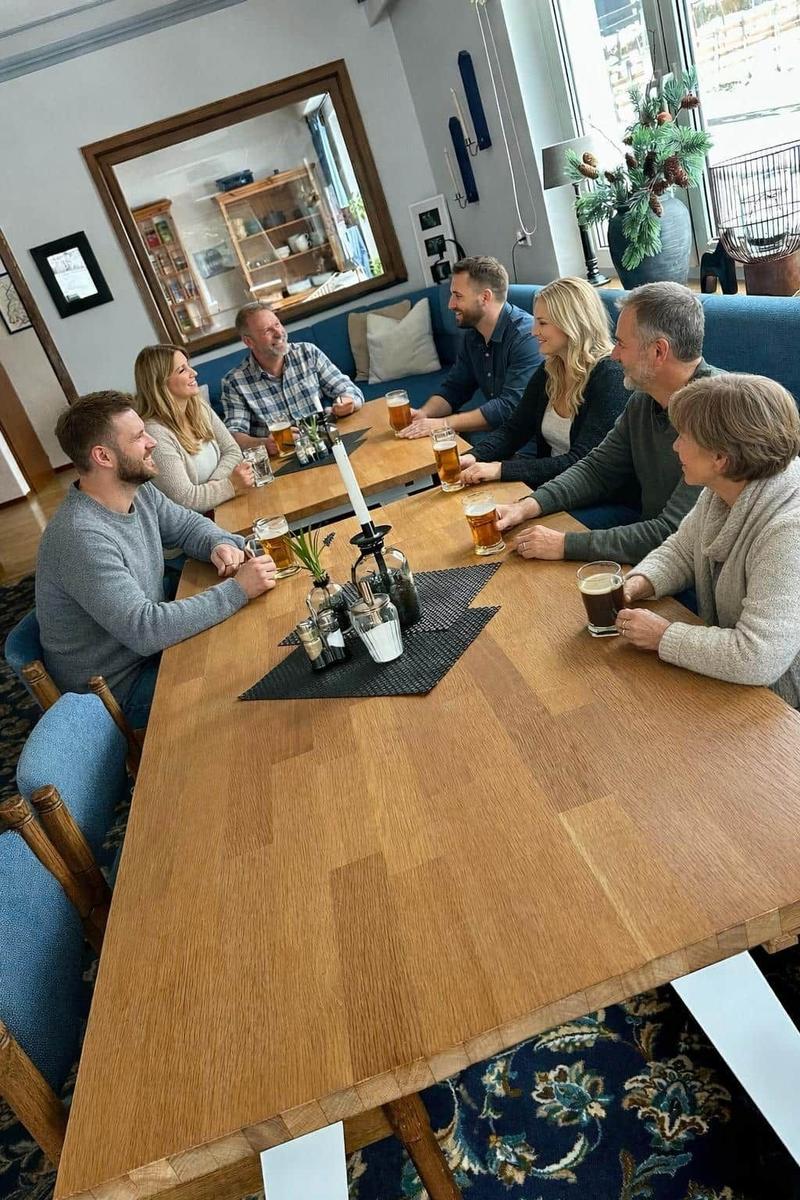 Gruppe sitzt um einen Holztisch mit Getränken. Blaue Sitzgelegenheiten und Teppich im Hintergrund.