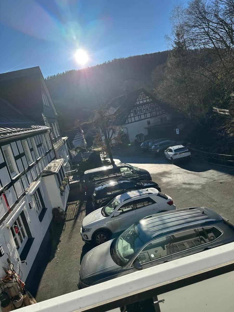 Parkplatz mit mehreren Autos vor traditionellen Häusern und bewaldeten Bergen unter Sonne.