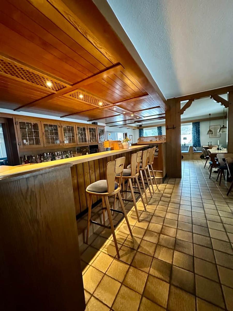 Bar mit Holzregalen und Sitzhöhenstühlen, Fliesenboden und Sicht auf Essbereich.