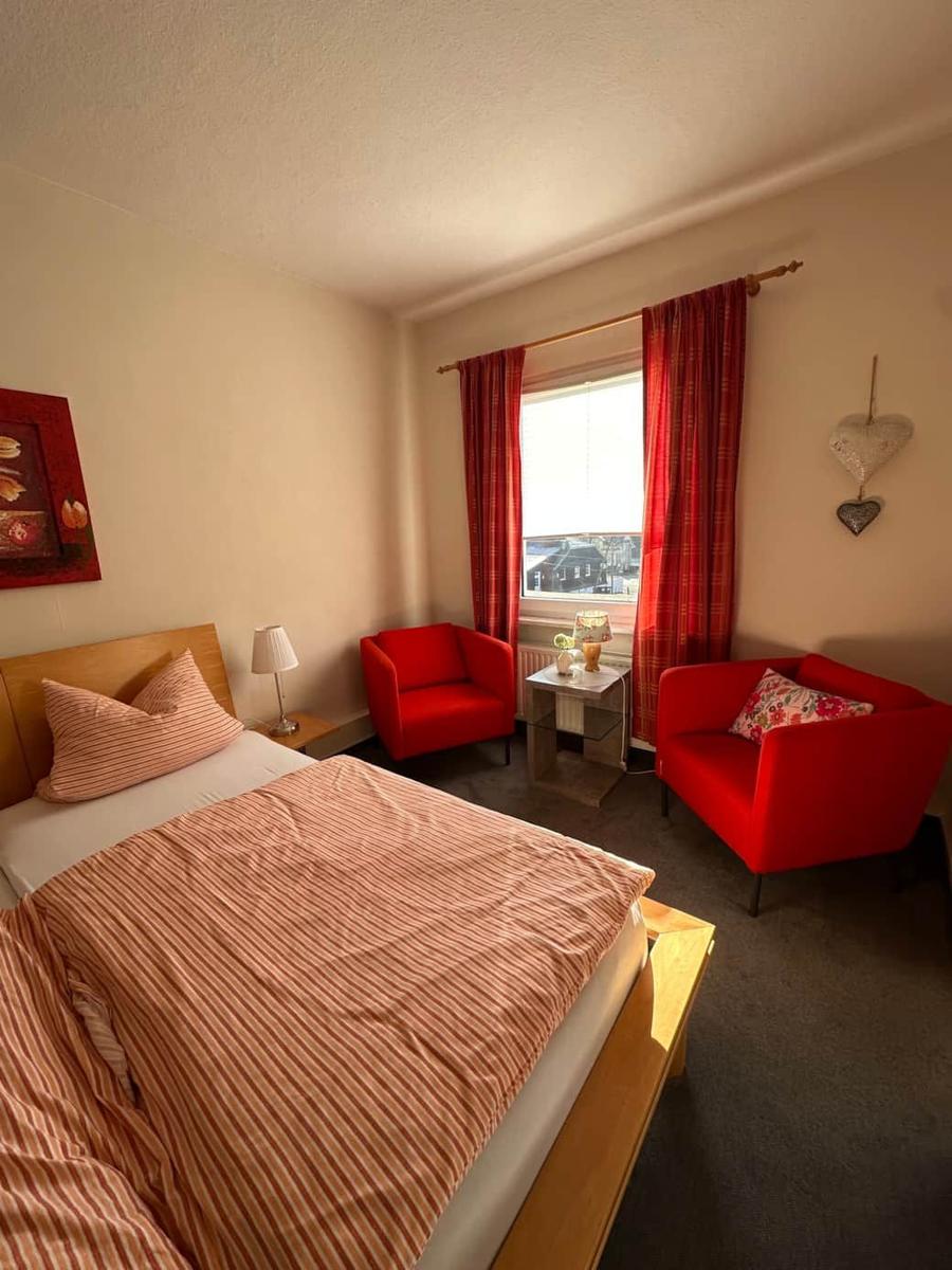 Ein Schlafzimmer mit Bett, zwei roten Sesseln und Fenster mit roten Vorhängen.