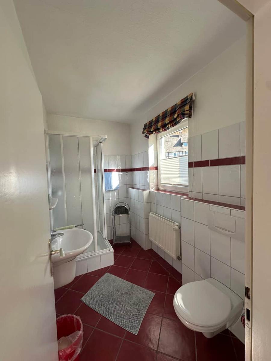 Badezimmer mit Dusche, Waschbecken und Toilette. Rote Fliesen und Fenster mit Vorhang.