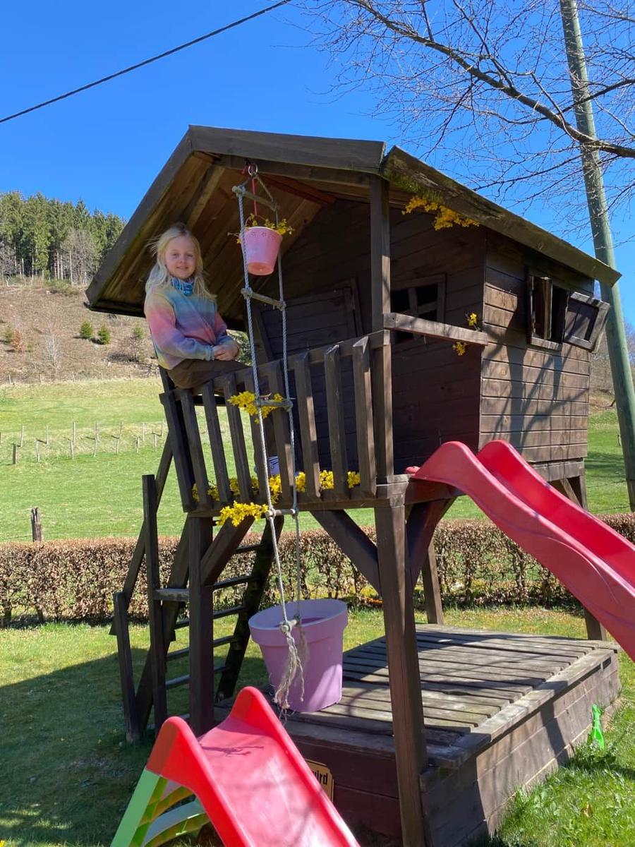 Ein Kind sitzt auf einer Holzspielburg mit roter Rutsche im Garten.