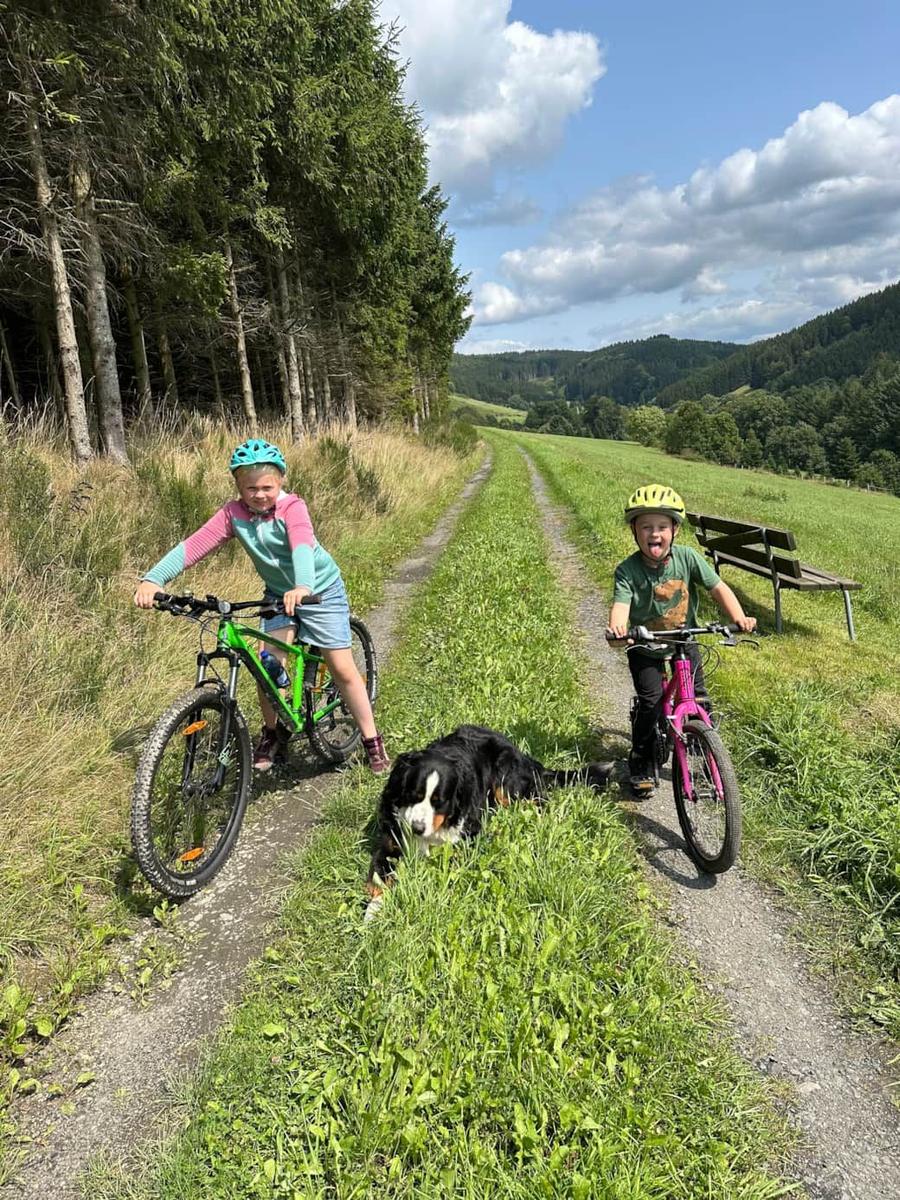 Zwei Kinder mit Fahrrädern und Hund auf Waldweg.