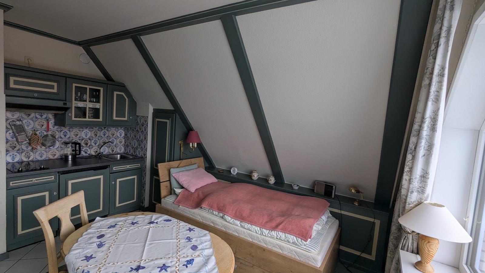 Ein Schlafzimmer mit kleiner Küche und Tisch. Holzbett mit rosa Bettwäsche.