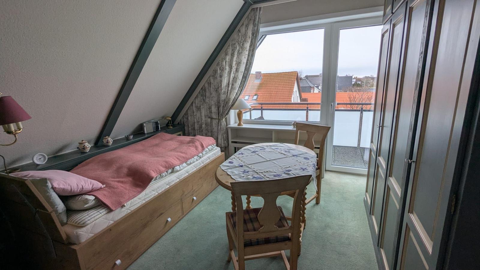 Schlafzimmer mit Dachschräge, Bett, Tisch und Balkonblick.
