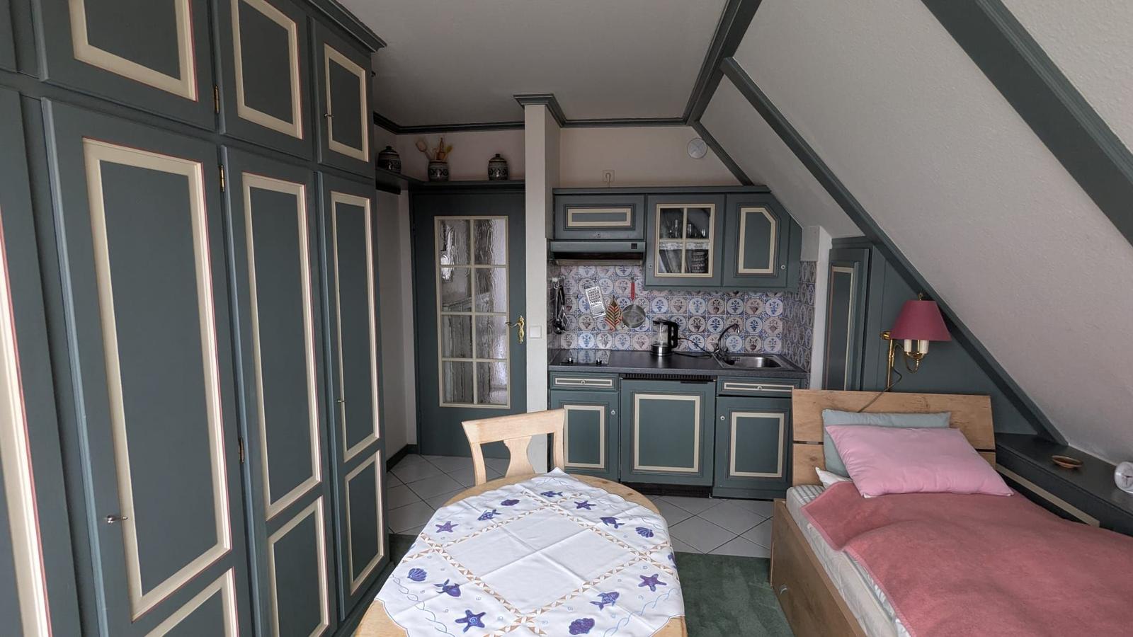 Kleines Zimmer mit Bett, Tisch und Küchenzeile unter Dachschrägen.