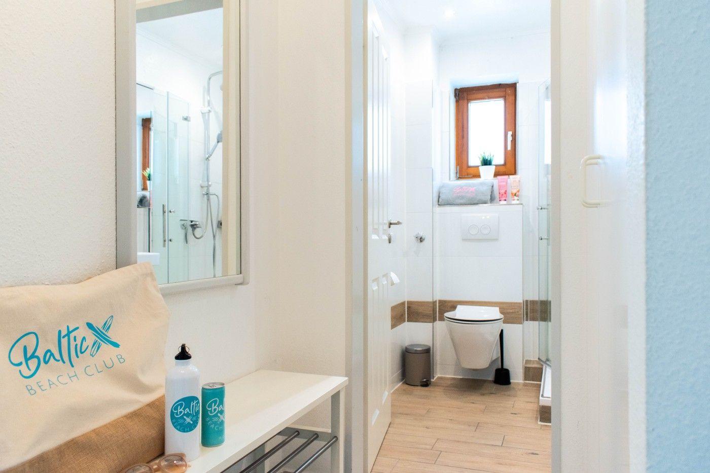 Badezimmer mit Dusche, Toilette und Spiegel. Auf Ablagefläche stehen Flasche und Dose.