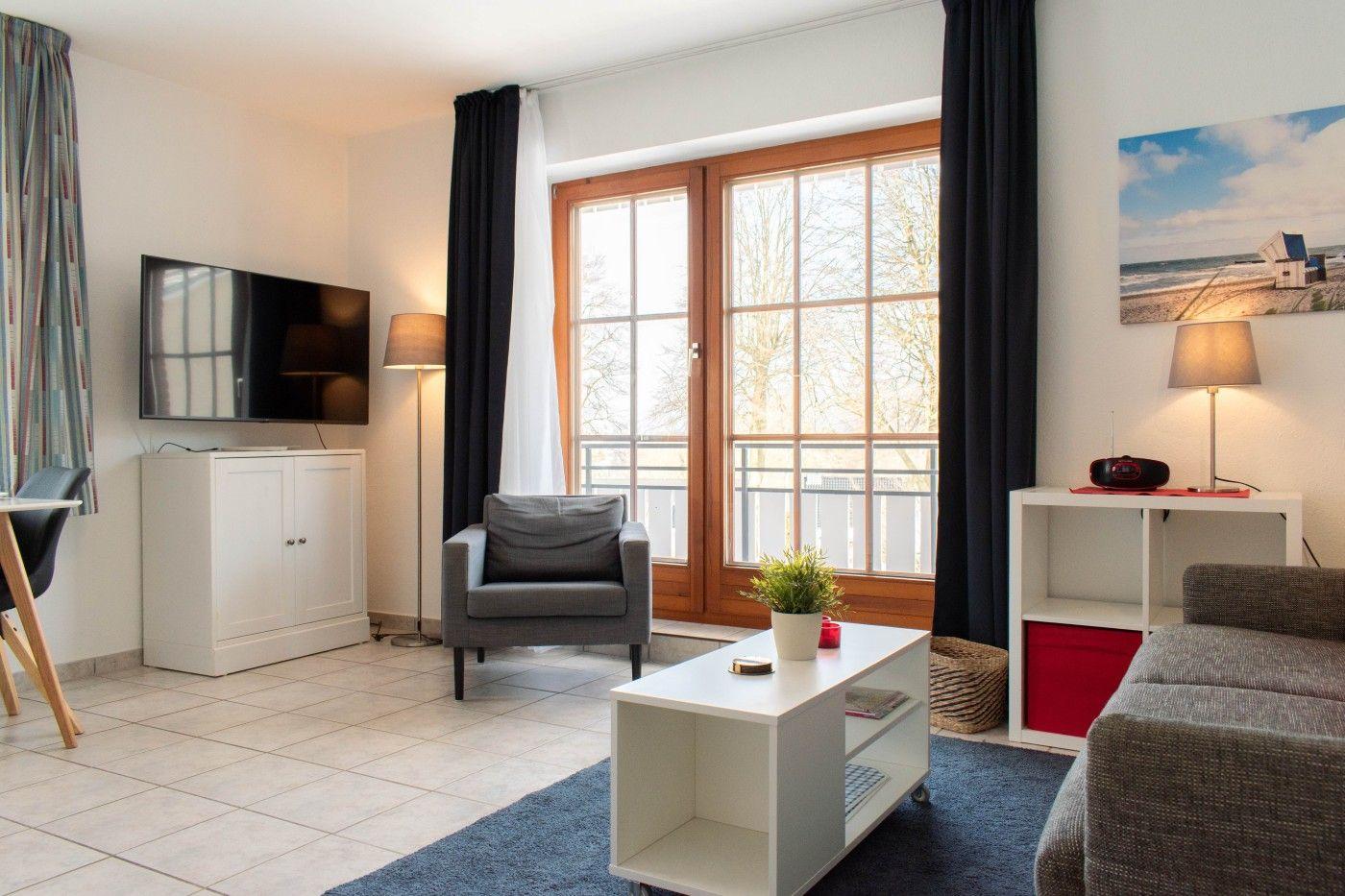 Wohnzimmer mit Sofa, Sessel, Fernseher und Balkonfenster.