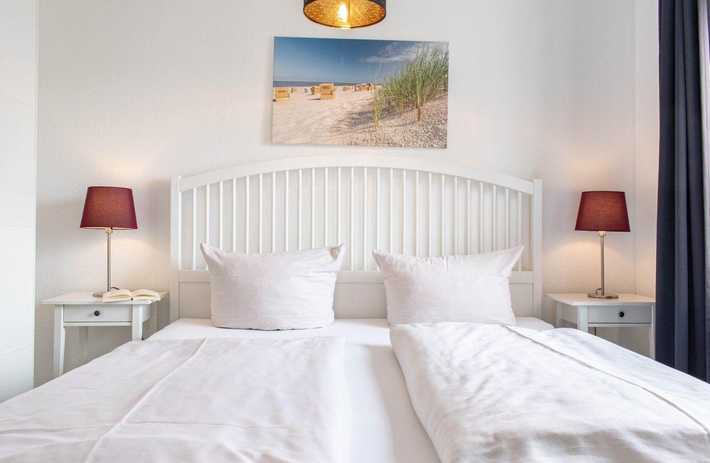 Schlafzimmer mit weißem Bett, zwei Nachttischen und einer Strandbilddekoration.