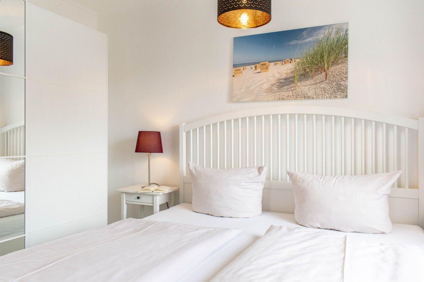 Schlafzimmer mit weißem Bett, Nachttisch und Wandbild. Spiegel und Deckenleuchte sichtbar.