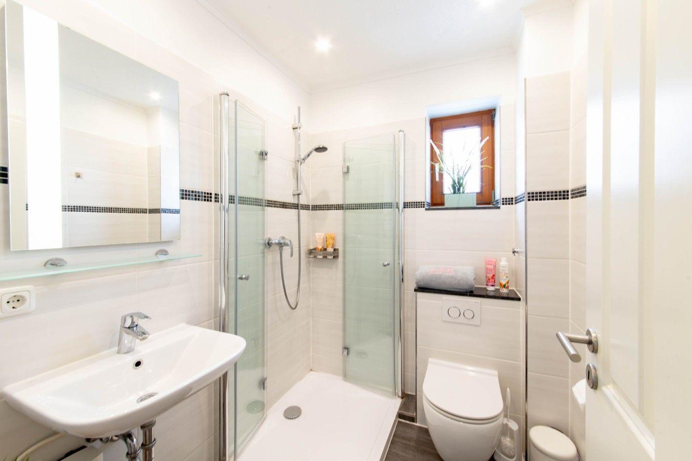 Badezimmer mit Dusche, Waschbecken und Toilette. Fenster mit Pflanze.