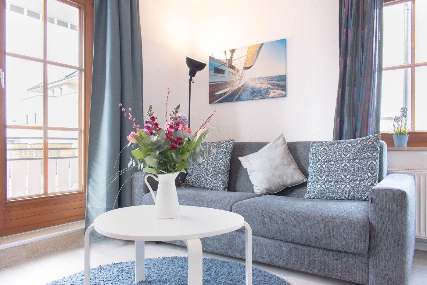 Ein grauer Sofa mit Kissen, weißer Tisch und Blumen vor Fenster.