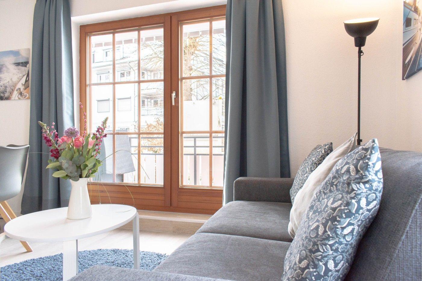 Ein Wohnzimmer mit Sofa, Tisch und Blumen vor Fenster mit Blick auf Balkon.