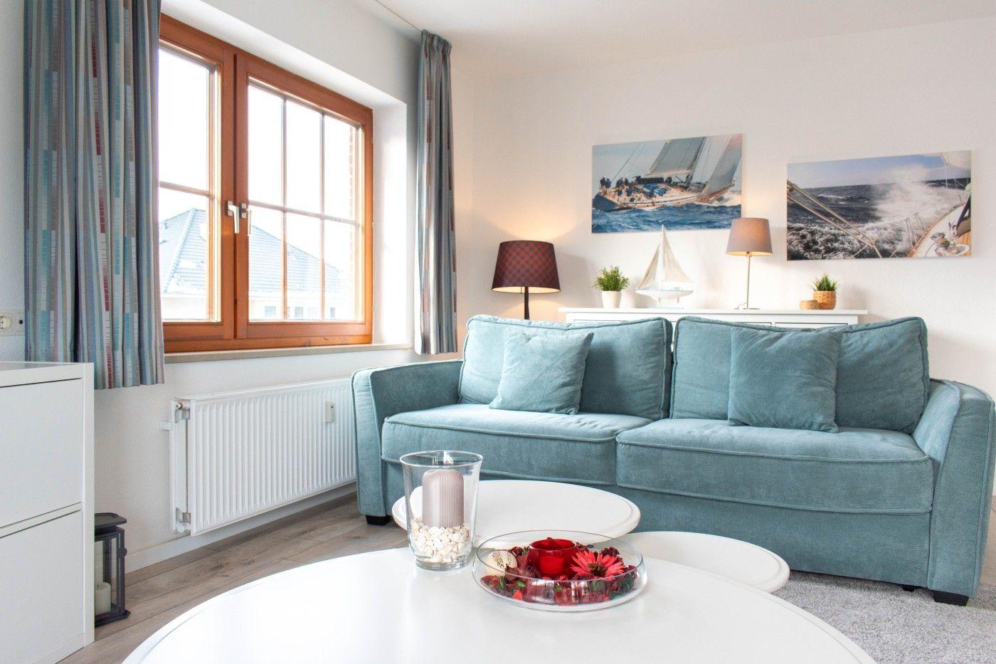 Ein Wohnzimmer mit blauem Sofa, weißen Tischen und Fenstern mit Vorhängen.