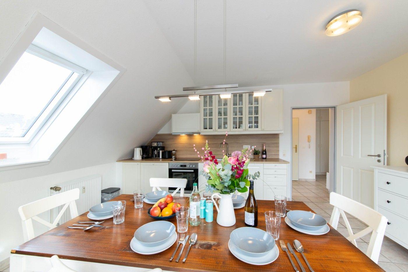 Esszimmer mit Holztisch, weißen Stühlen und Küchenbereich unter Dachschrägen.