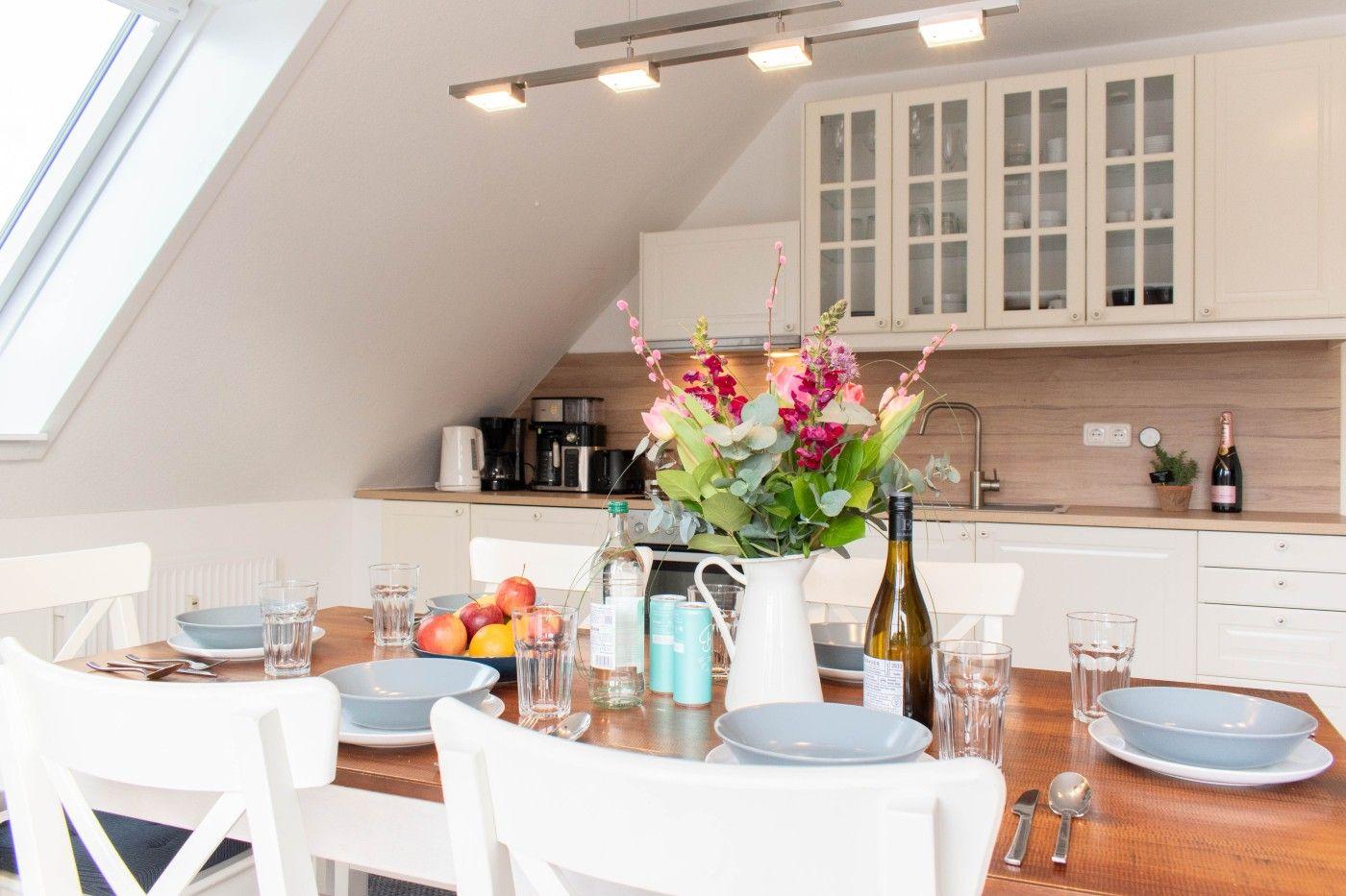 Essbereich mit Tisch, Stühlen und Blumenarrangement. Küche mit Schränken und Arbeitsfläche.