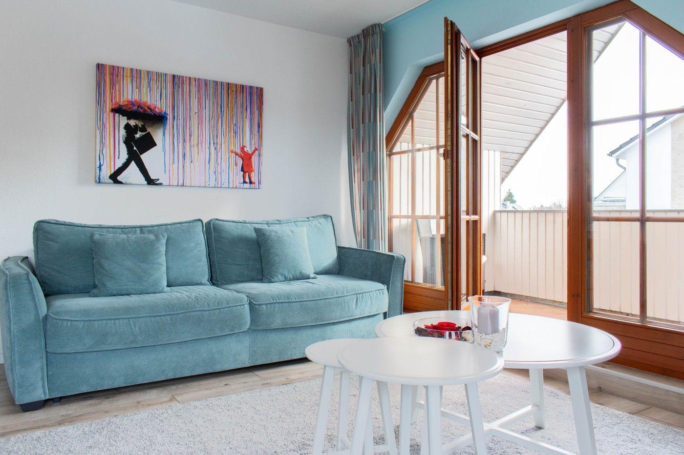 Ein Wohnzimmer mit blauem Sofa, weißen Tischen und einer großen Fensterfront.