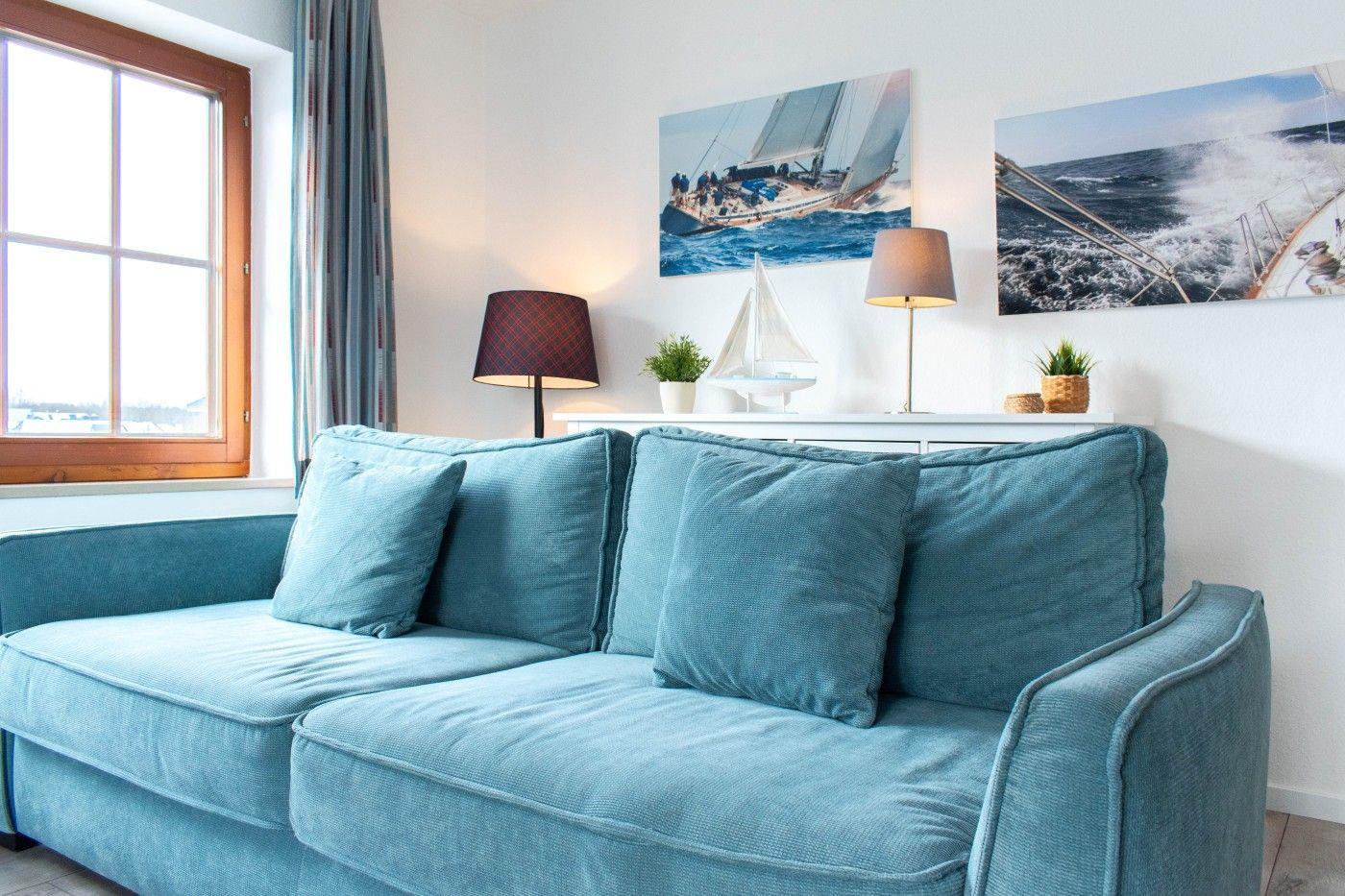 Blauer Sofa mit Kissen, Wandbilder von Segelbooten und Fenster mit Holzrahmen.