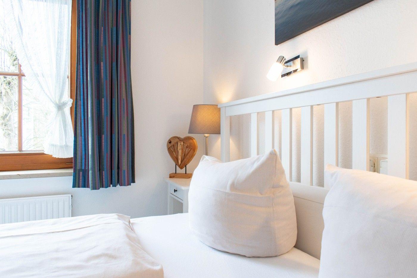 Schlafzimmer mit weißem Bett, Nachttisch, Lampe und Fenster mit Vorhängen.