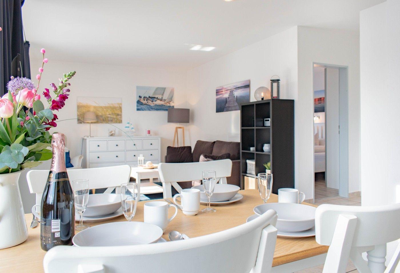 Essbereich mit Tisch und Stühlen, Blumen und Flasche. Im Hintergrund Wohnzimmer und Schlafzimmer.