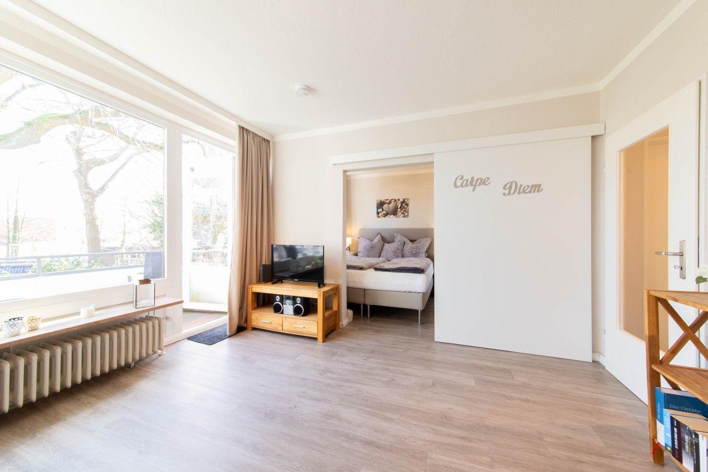 Wohnzimmer mit Schlafzimmer dahinter. Große Fenster, Holzboden und TV.