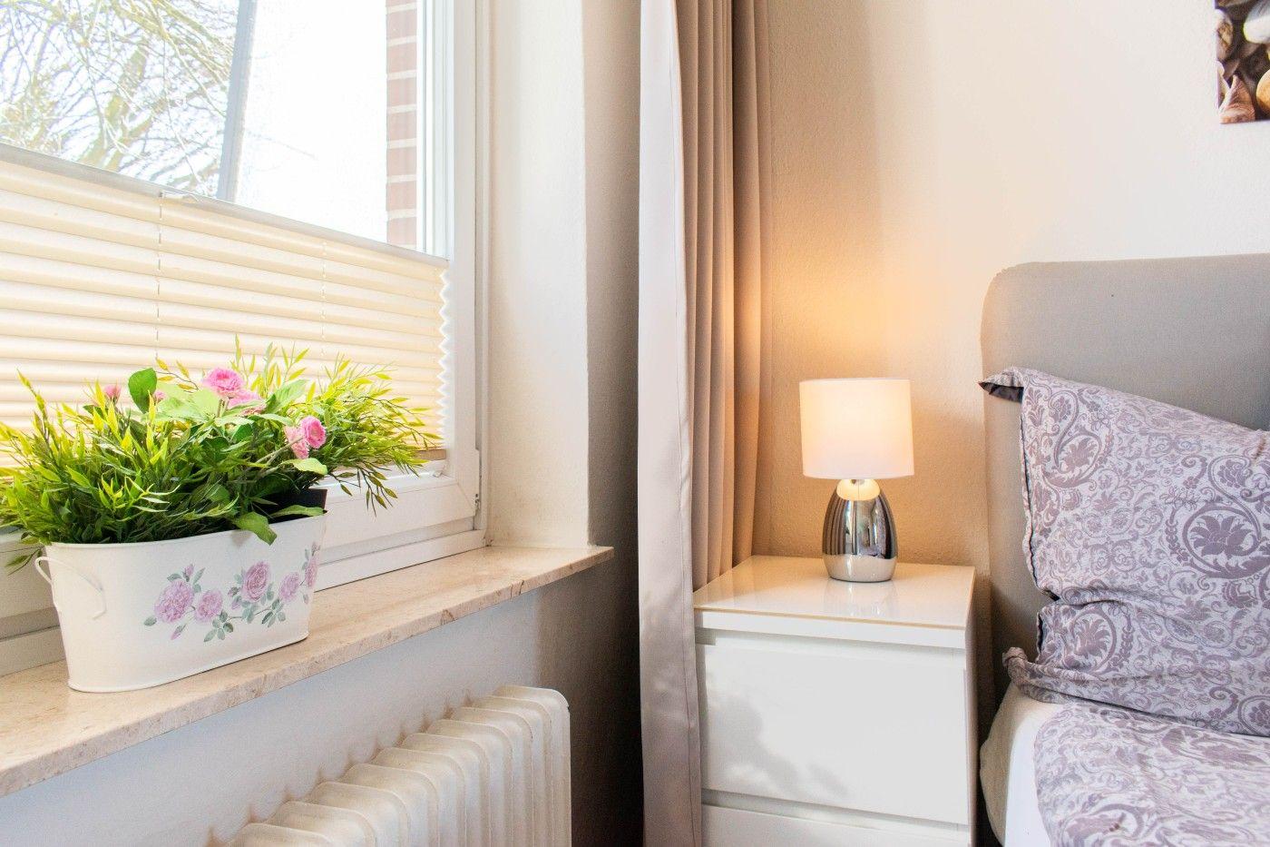 Schlafzimmer mit Bett, Nachttisch und Pflanze am Fenster.