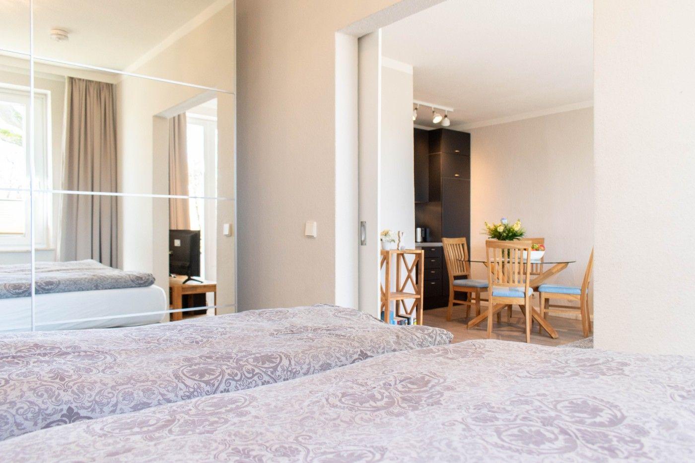 Zwei Betten mit gemusterten Bettwäsche. Im Hintergrund ist Esszimmer mit Holztisch und Stühlen.