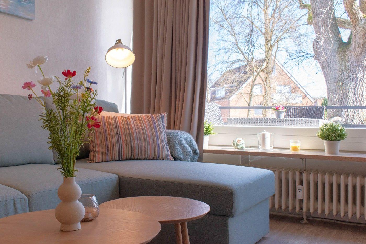 Wohnzimmer mit Sofa, Tisch und Blumen. Blick auf Balkon mit Pflanzen und Baum.
