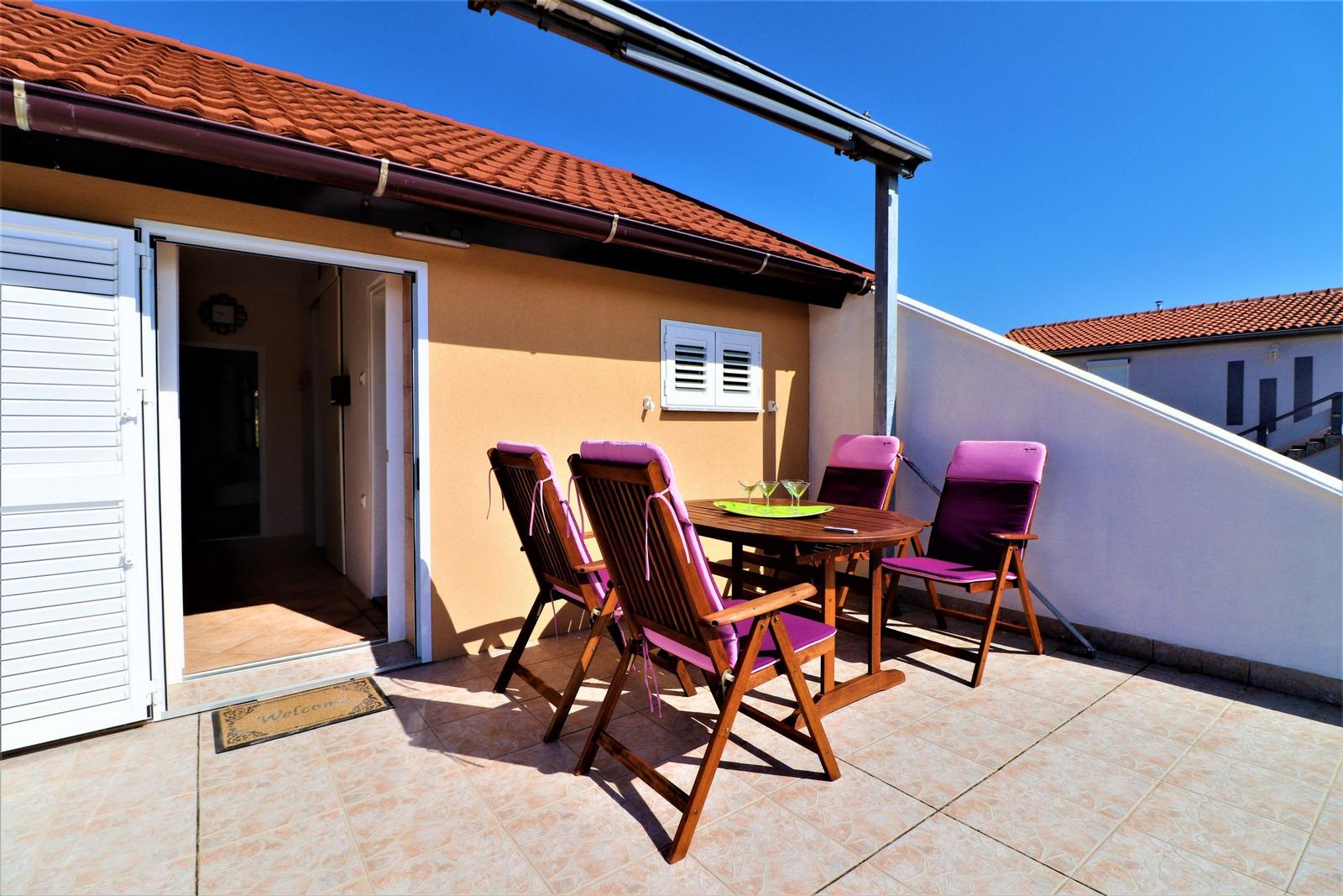 Terrasse mit Holztisch und vier Stühlen mit lila Sitzkissen. Tür führt ins Haus. Hintergrund: Haus mit rotem Dach.