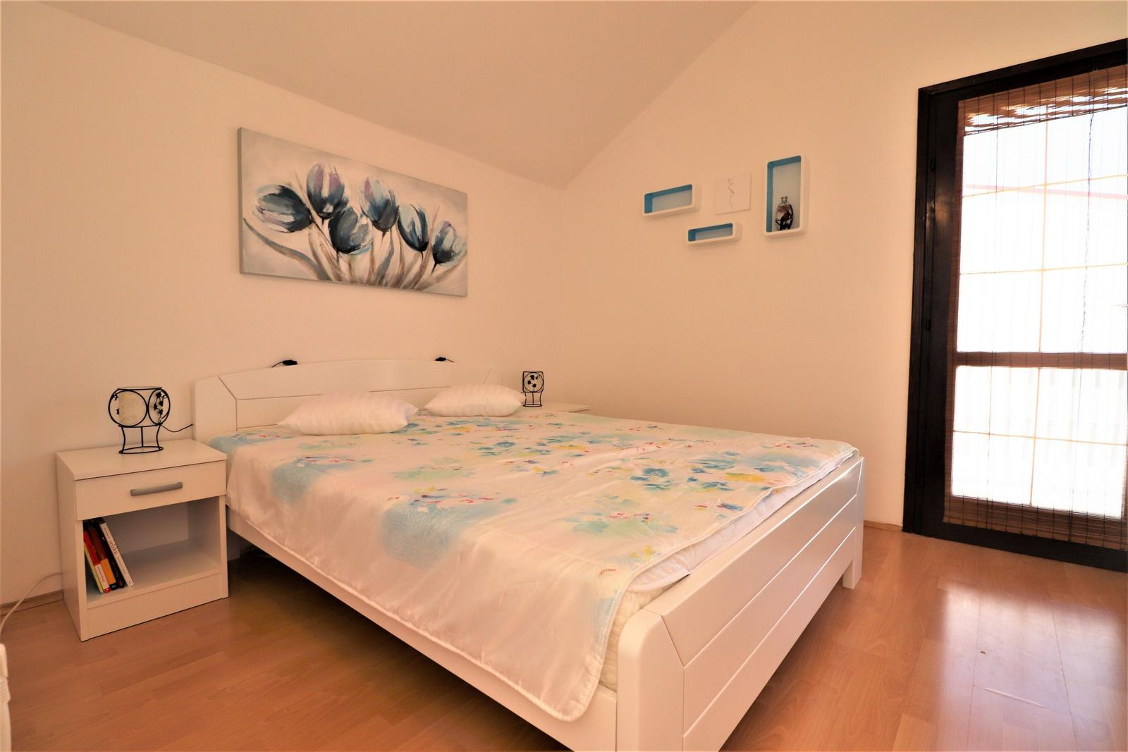 Schlafzimmer mit weißem Bett, Holzboden und Fenster mit Jalousie.