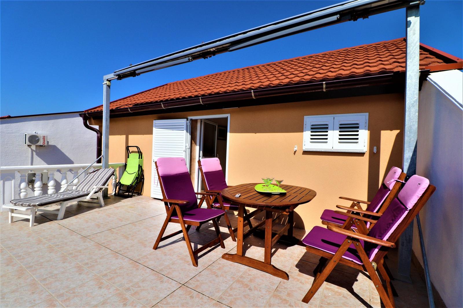 Terrasse mit Tisch, vier Stühlen und Liegestuhl unter Sonnenschutz.