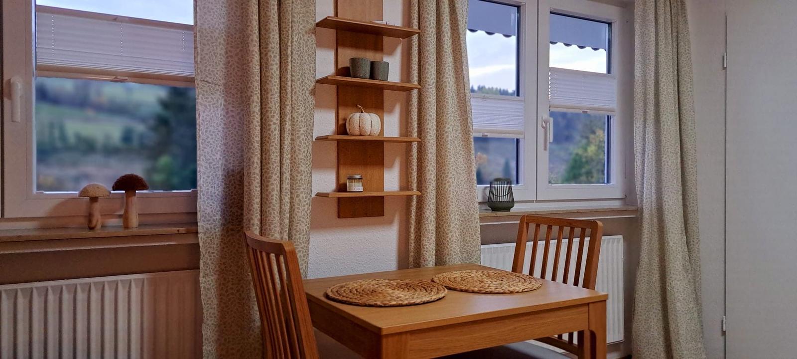 Holztisch mit zwei Stühlen, Wandregal und Fenster mit Blick auf Landschaft.