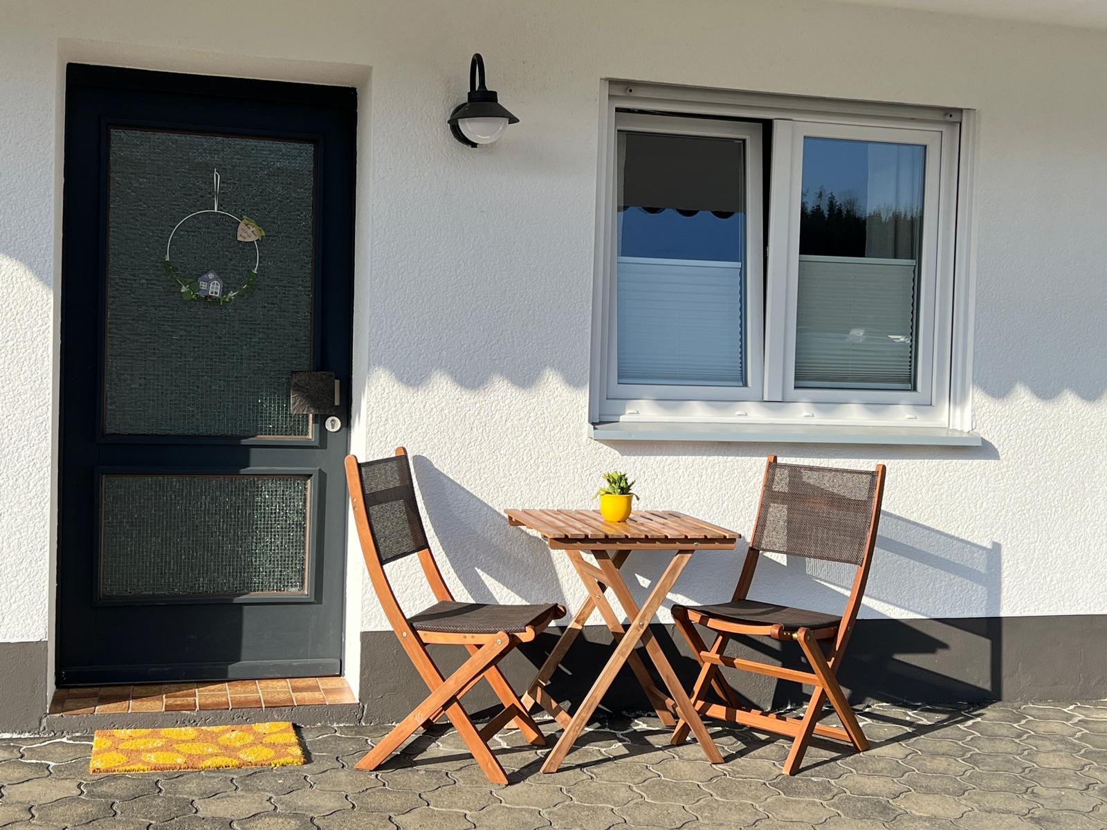 Vor dem Eingang steht ein Tisch mit zwei Stühlen. Eine Tür und ein Fenster sind sichtbar.