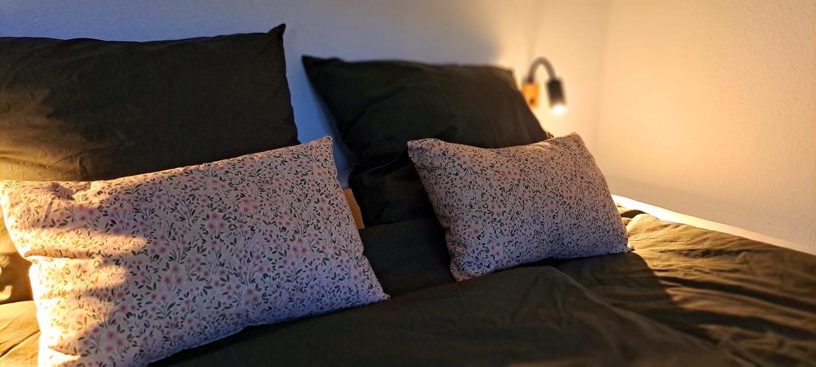 Bettkopfteil mit zwei Kissen und zwei Kissen mit Blumenmuster. Wandlampe an der Wand.