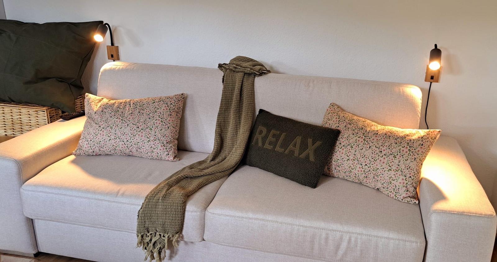 Ein Sofa mit Kissen und einer Decke, an den Wänden hängen Wandlampen.