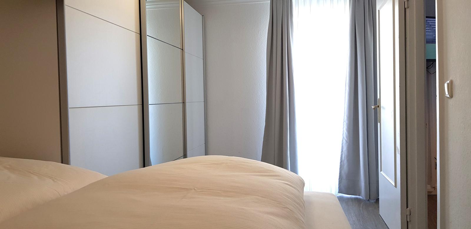 Schlafzimmer mit Bett, Kleiderschrank und Fenster mit Vorhängen.